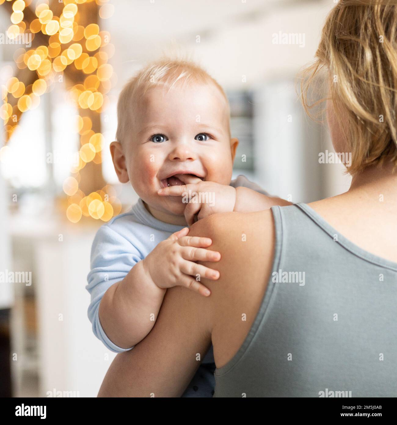 Blonde Mutter hält ihren niedlichen, fröhlichen kleinen Jungen. Süßes Kleinkind, das in die Kamera schaut und zu Hause lächelt Stockfoto
