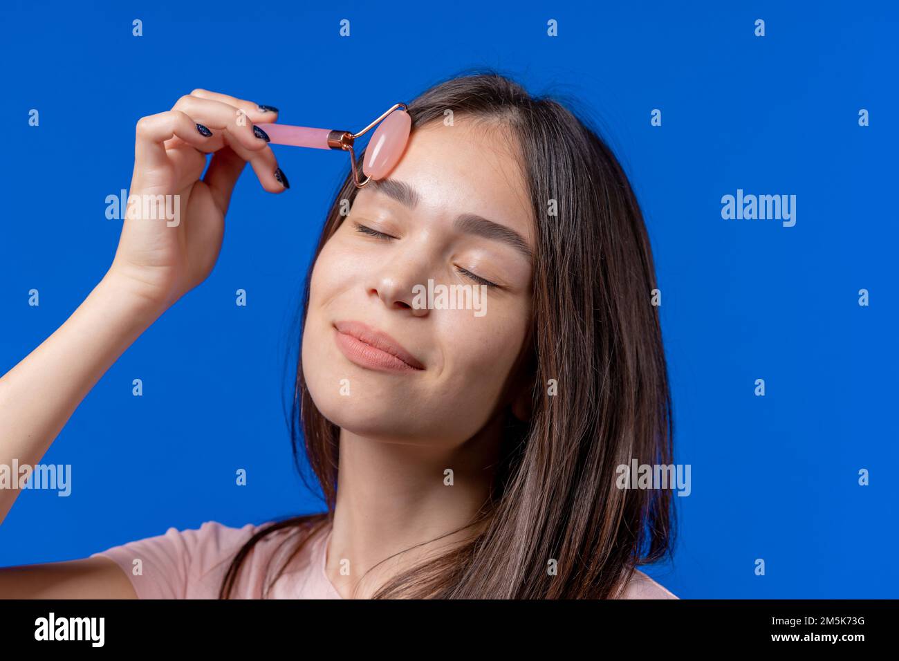Junge Frau, die eine Gesichtsmassage mit Rosenquarzwalze auf blauem Hintergrund macht. Gesichtsproflege, Schönheitsrituale, Kosmetik, Anti-Aging und Anti Stockfoto
