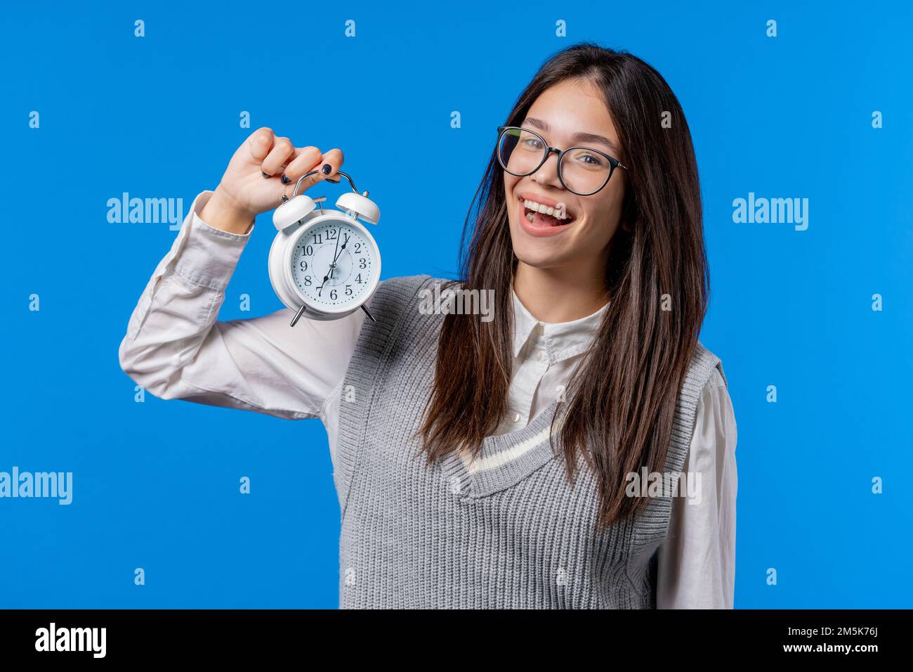Glücklicher asiatischer Schüler Teenager mit Wecker auf blauem Hintergrund. Am frühen Morgen, korrekte Tagesroutine, im Moment leben, jede Minute schätzen Stockfoto