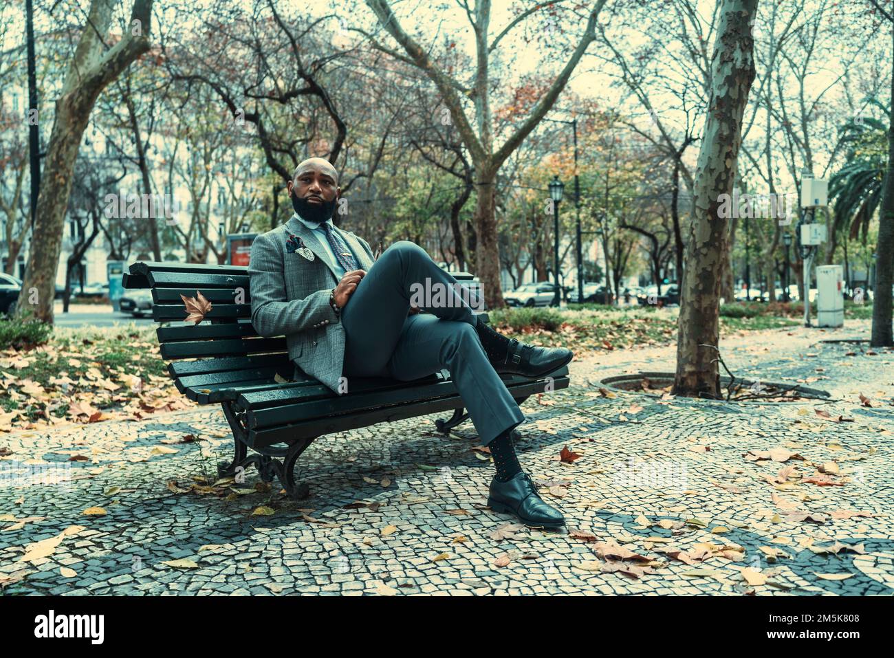 Ein gut aussehender, bärtiger, schwarzer Unternehmer in einem eleganten Outfit, der auf einer hölzernen Straßenbank auf dem Bürgersteig sitzt; ein afroamerikanischer Busin Stockfoto