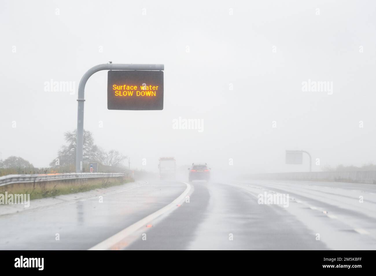 Veränderliches Hinweisschild „VERLANGSAMUNG durch Oberflächenwasser“ auf der britischen Autobahn bei starkem Regen Stockfoto