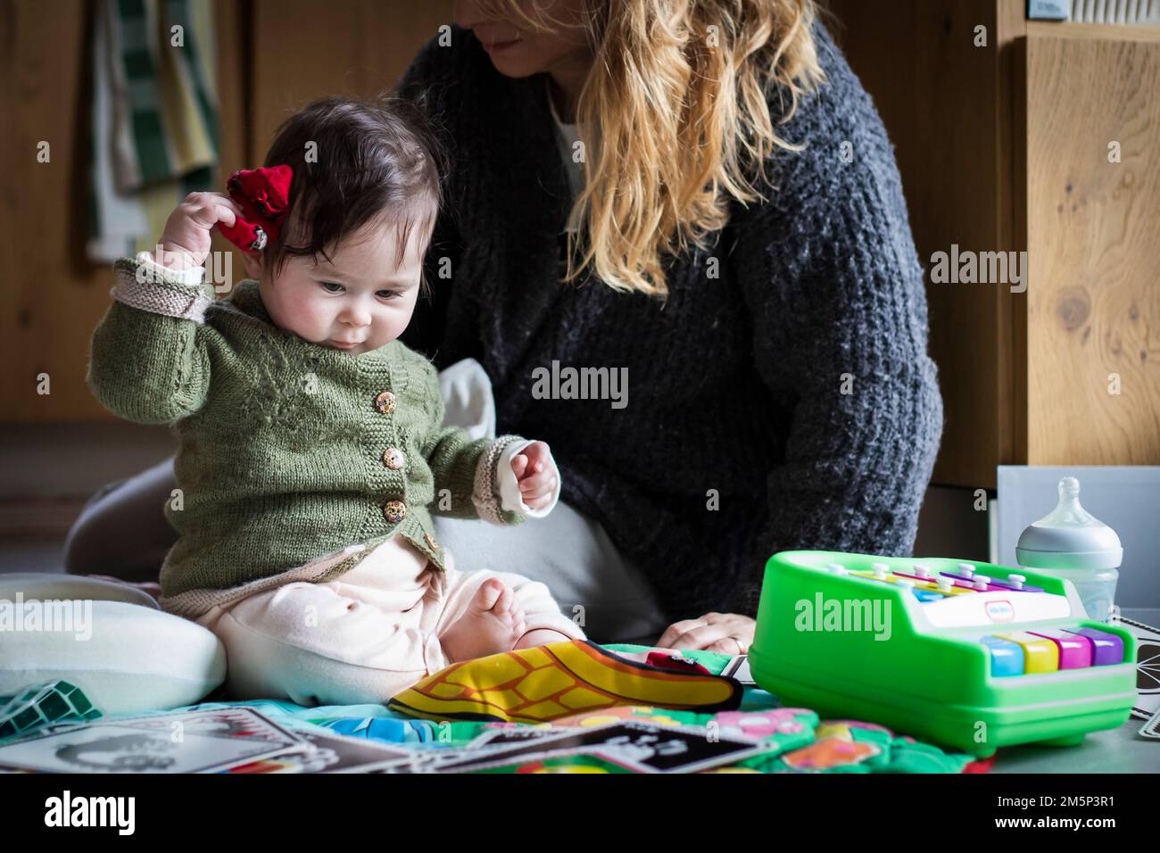 Das niedliche Baby spielt auf einem Spielvorleger neben seiner Mutter und hält ihre roten Socken statt ihrer Spielzeuge vor sich. Echte Umgebung für Zuhause Stockfoto