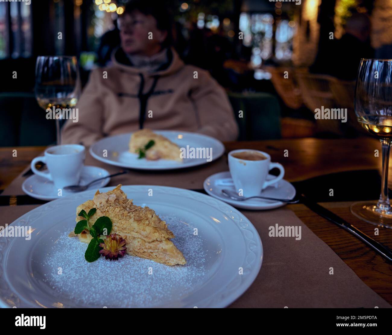 Napoleonkuchen auf einem weißen Keramikteller und Kaffeetassen, Gläser mit unvollendetem Weißwein und eine ziemlich reife Frau, die im Bac aus dem Fenster schaut Stockfoto