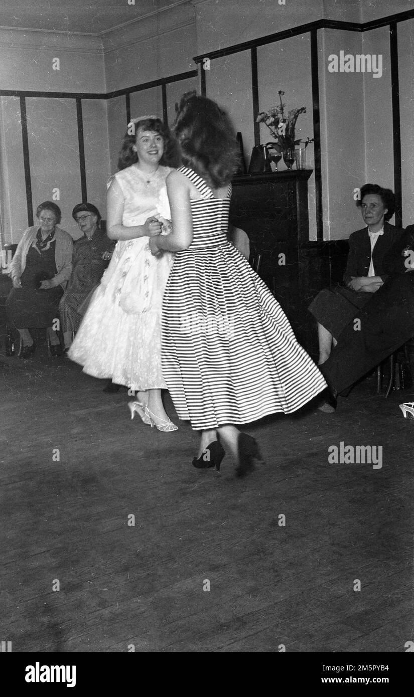 1950er Jahre, historisch, zwei junge Damen in langen Kleidern und High Heels tanzen zusammen auf dem Holzboden eines Zimmers bei einem Hochzeitsempfang in England, Großbritannien. Andere Gäste, die neben der Beobachtung sitzen. Stockfoto