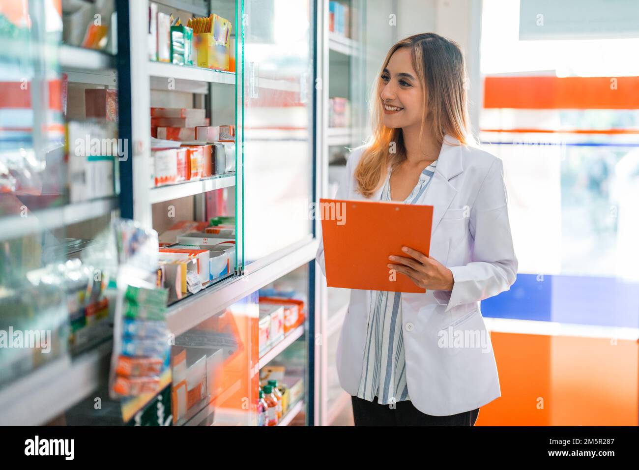 Schöne Apothekerin in Uniform, die mit dem Klemmbrett in der Apotheke arbeitet Stockfoto