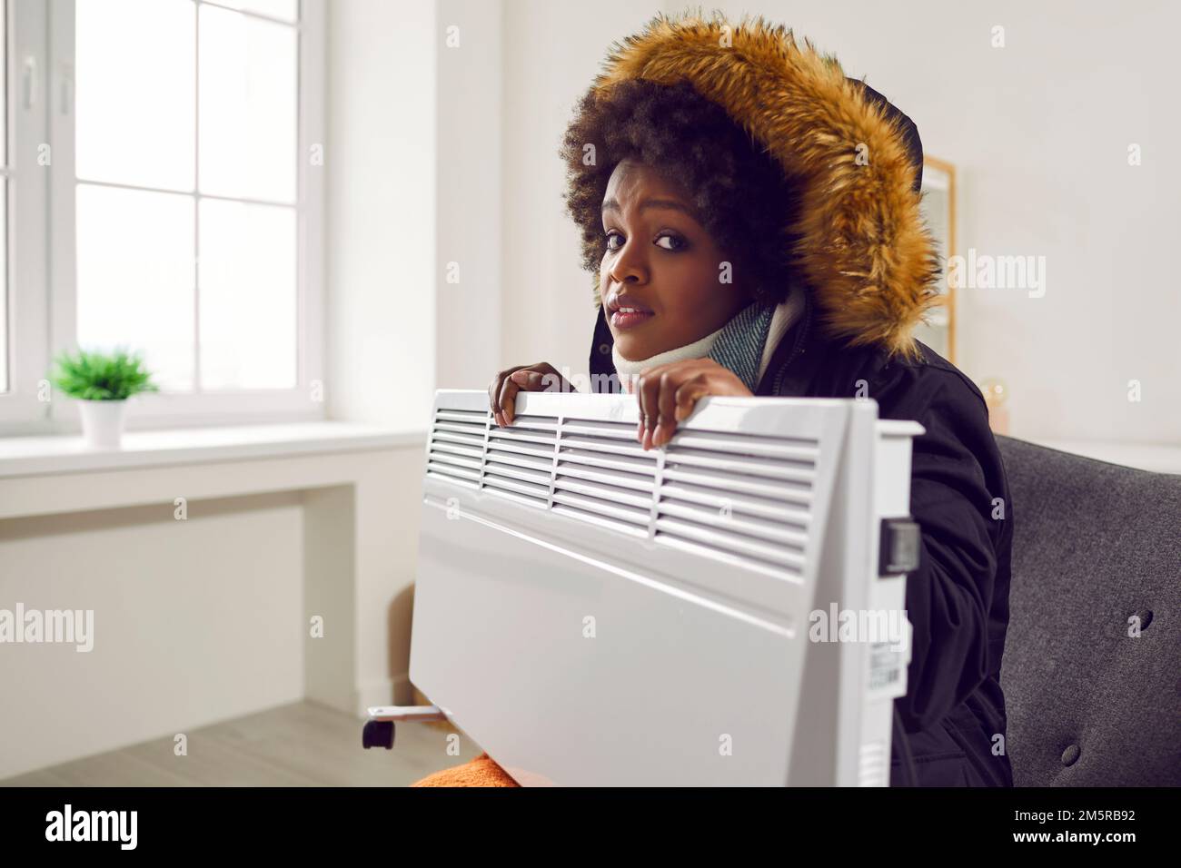 Eine afroamerikanische Frau in Winterkleidung, die auf der Couch sitzt, mit elektrischer Heizung im Kühlhaus Stockfoto