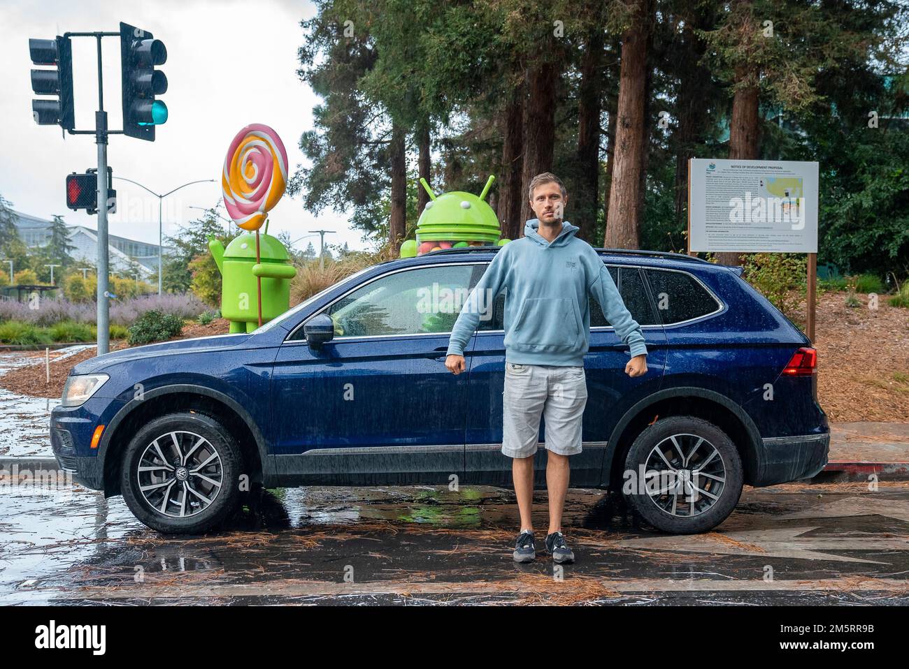 Ein Mann vor einem blauen Auto auf einer nassen Straße mit android-Statuen im Hintergrund Stockfoto