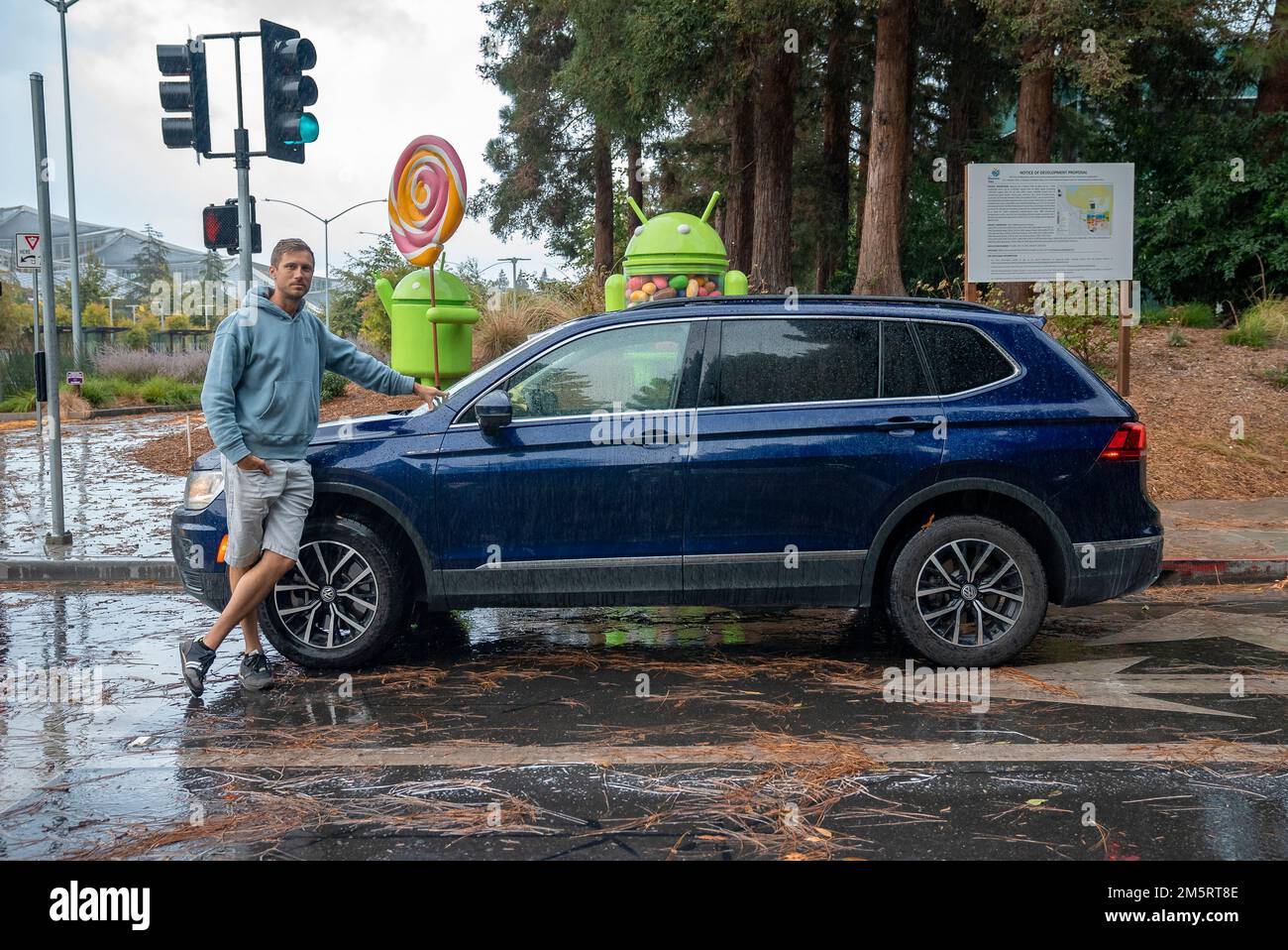Ein Mann, der sich an einem blauen Auto lehnt und auf einer nassen Straße vor android-Statuen parkt Stockfoto