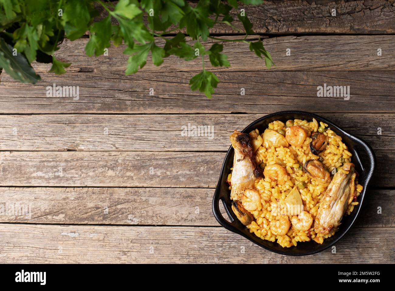 Schwarzer Teller mit gemischter Paella, Fleisch und Fisch, mit rustikalem Holzhintergrund und Petersilienzweigen, Kopierbereich. Draufsicht. Stockfoto