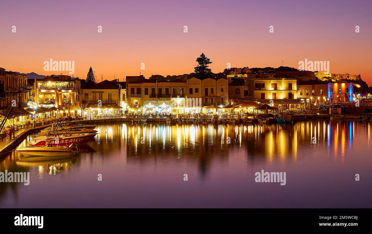 Venezianischer Hafen, Dämmerung, Nachtaufnahmen, orangefarbener Himmel, bunte Reihen von Häusern, Wasserreflexion, künstliches Licht, Boote, Rethimnon, Zentrum von Kreta Stockfoto