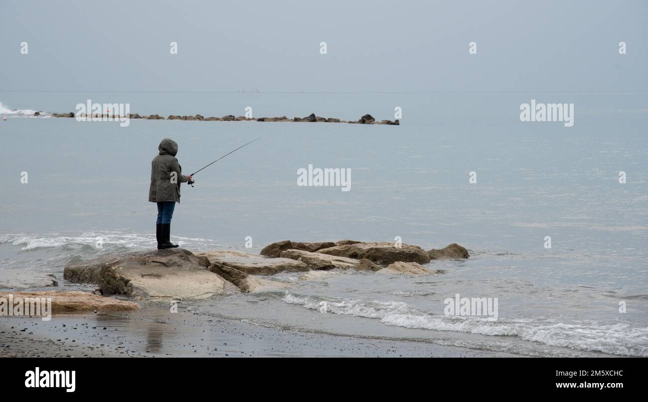 Unbekannte Person, die im Winter auf Felsen steht und mit Rote auf dem Meer angeln Stockfoto