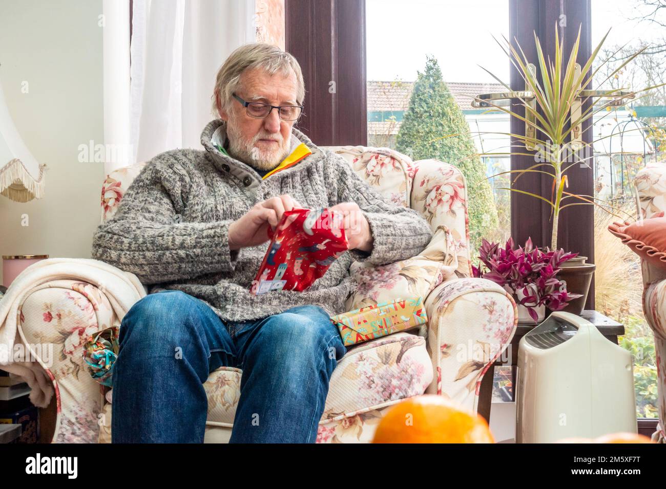 Ein Mann öffnet ein Weihnachtsgeschenk, während er in einem bequemen Sessel saß. Stockfoto
