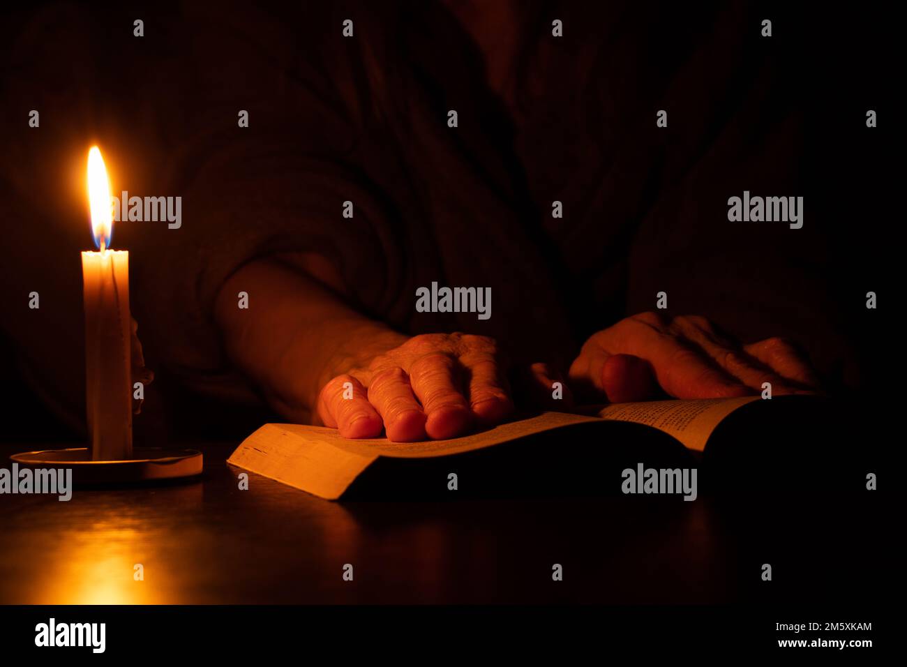 Großmutters Hände liegen auf einem offenen Buch auf dem Tisch, am Licht einer Kerze im Dunkeln, und lesen ein Gebet mit einer Kerze Stockfoto