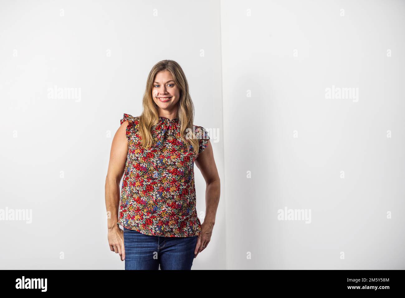 Eine wunderschöne weiße Frau in den Vierzigern in einem lässigen Oberteil mit Blumenmuster und Jeans, die an einer weißen Wand mit Kopierbereich stehen Stockfoto