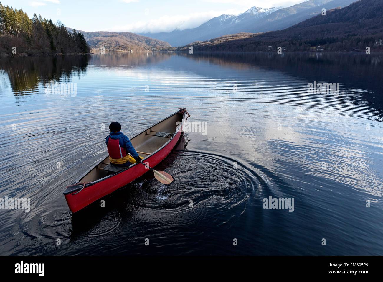 Einheimisches Kind paddelt in einem roten Kanu an einem kalten Wintertag auf einem ruhigen und schönen See Bohinj in Gorenjska in Slowenien Stockfoto