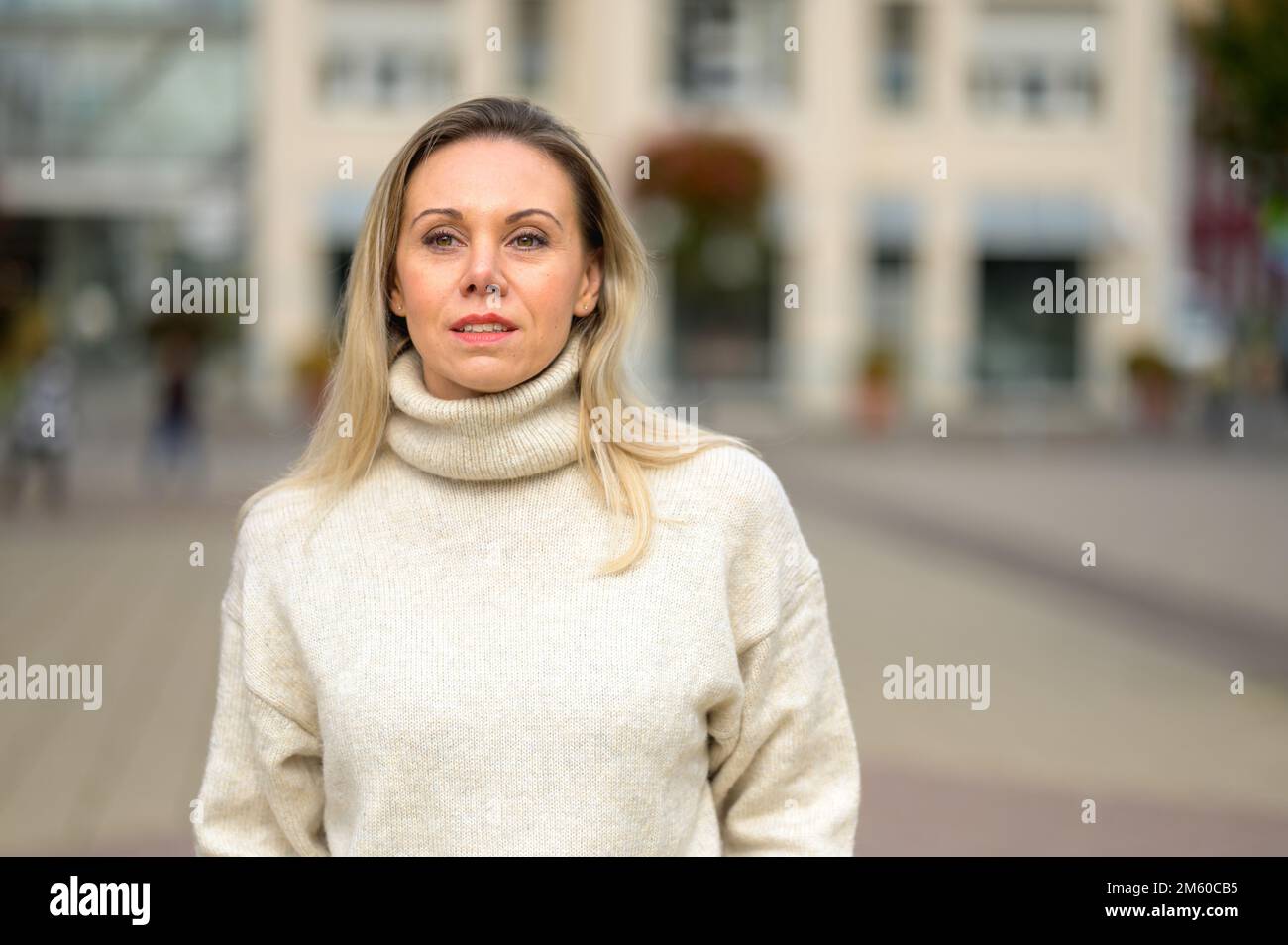 Attraktive blonde Frau mittleren Alters, die durch eine urbane Umgebung mit einem durchdachten Blick in die Ferne geht Stockfoto