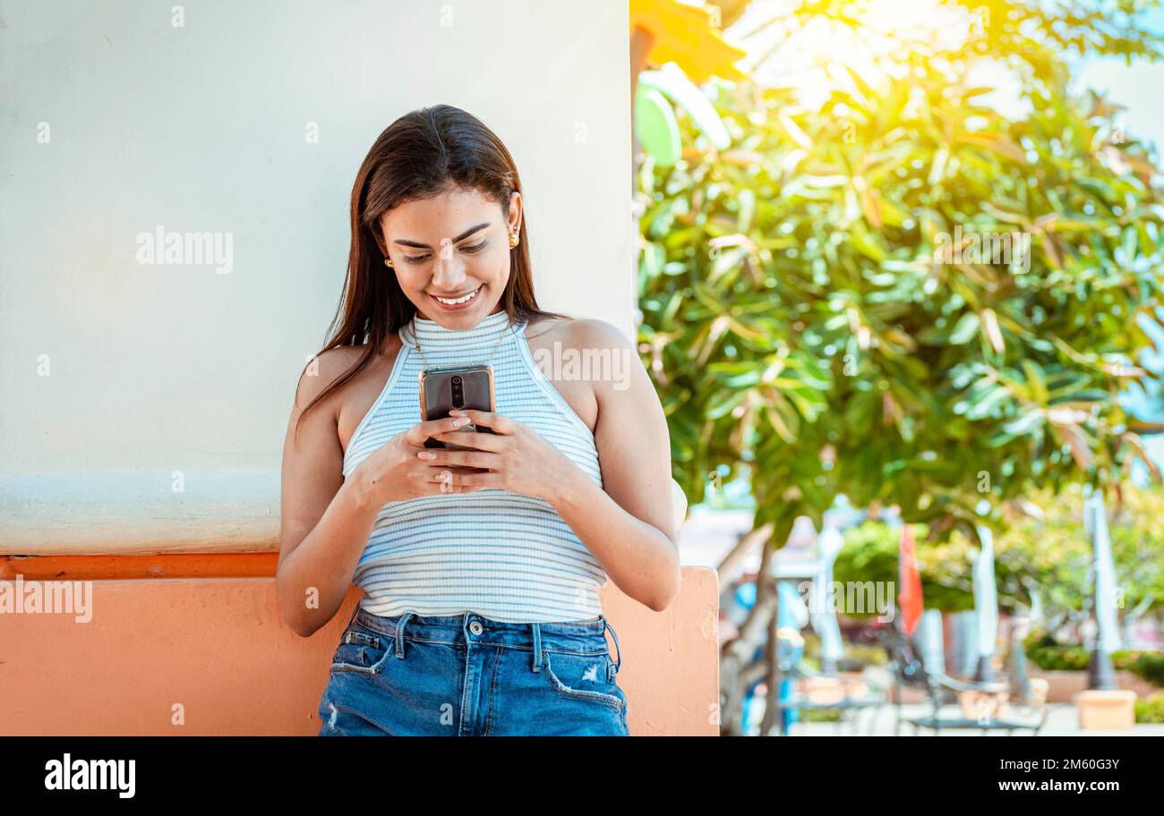 Glückliches Mädchen mit Handy, das gegen eine Wand lehnt. Ein junges Mädchen lehnt sich an eine Wand, ihr Handy lächelt, eine junge Frau, die SMS auf dem Handy schreibt Stockfoto