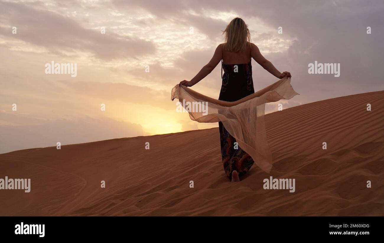Frau in Kleid mit Seidenschal, Blick auf Wüstensand bei Sonnenuntergang. Querformat. Schönheit, Mode und Reisekonzept Stockfoto