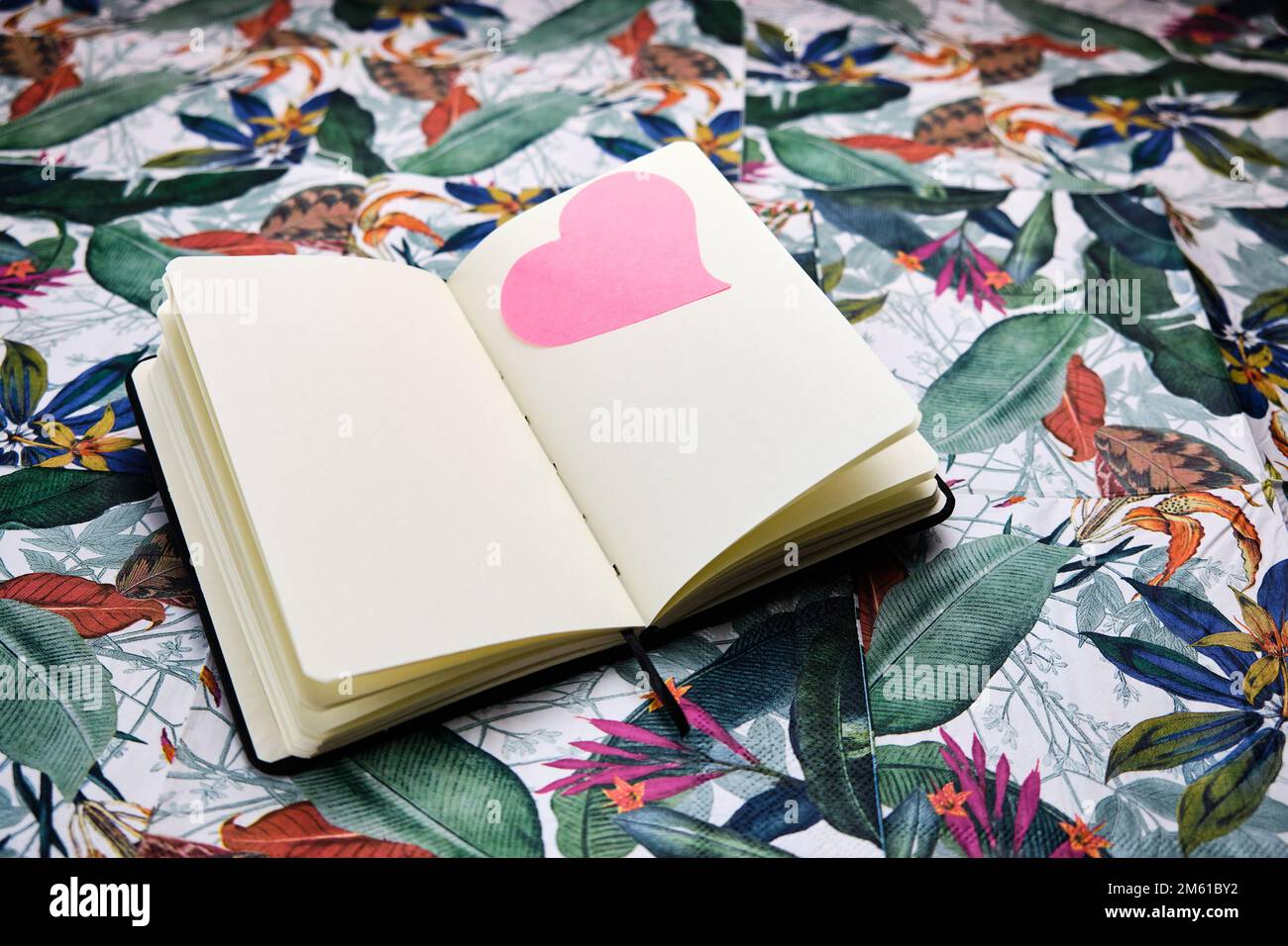 Das Notizbuch wird zu einem leeren Blatt mit einem rosa Herz auf einem Laken geöffnet, auf einem Tisch mit einem tropischen Muster. Stockfoto