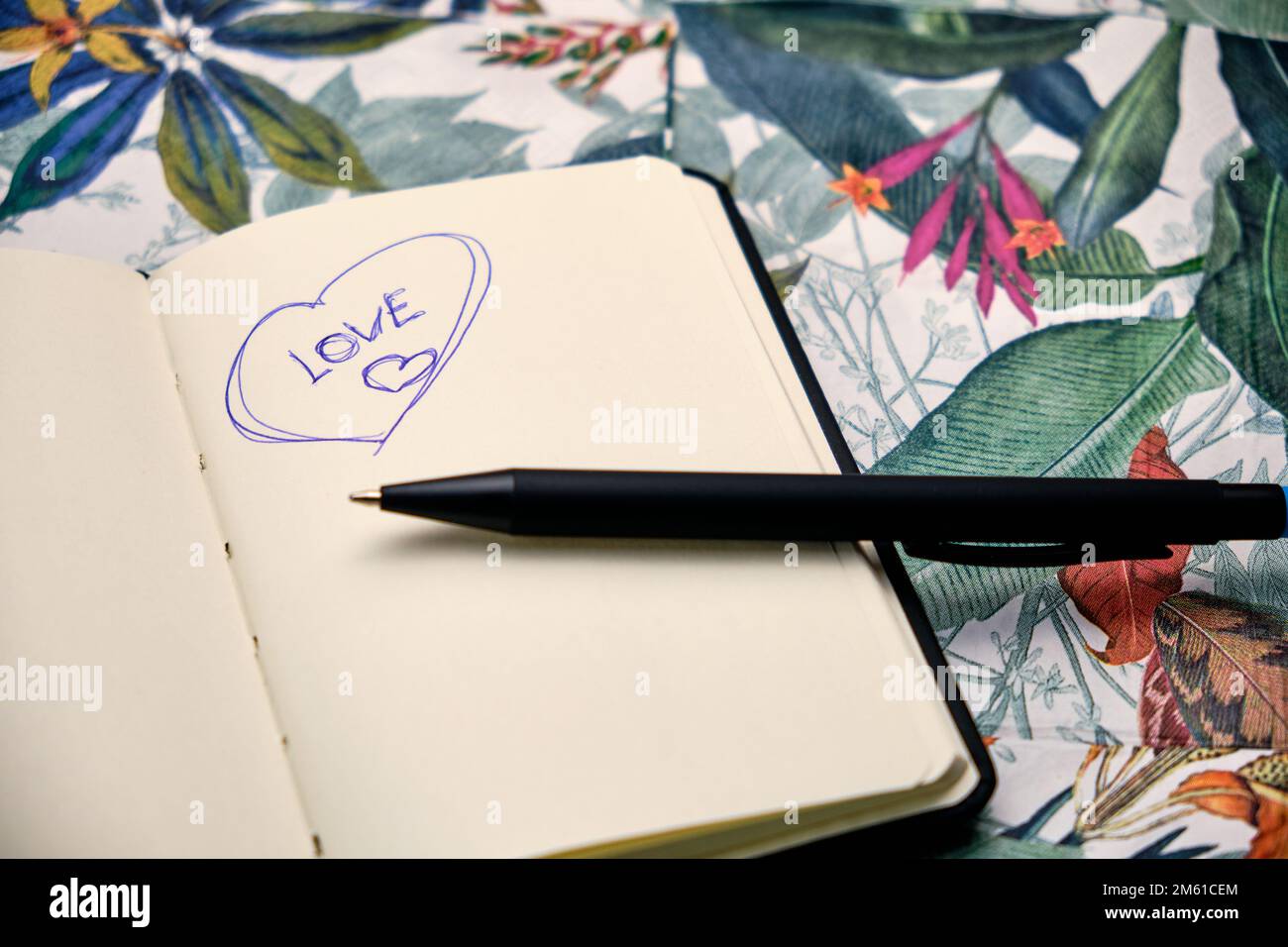 Öffnen Sie das Notizbuch mit einem Stift und schreiben Sie das Wort "Liebe" auf einen Tisch mit einem tropischen Muster. Stockfoto