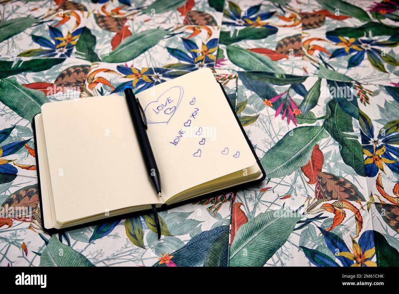 Öffne das Tagebuch mit einem Stift und schreibe das Wort Liebe auf einen Tisch mit einem tropischen Muster. Stockfoto