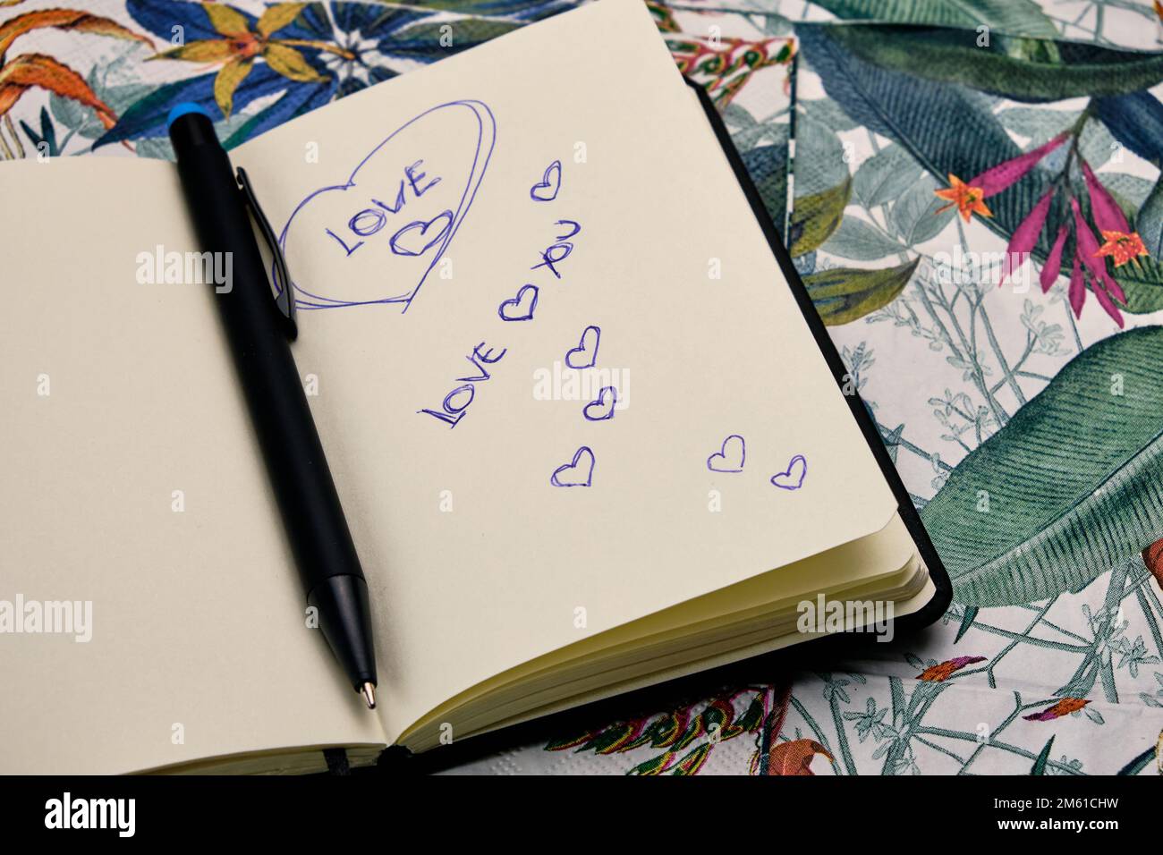 Öffne das Tagebuch mit einem Stift und schreibe das Wort Liebe auf einen Tisch mit einem tropischen Muster. Stockfoto