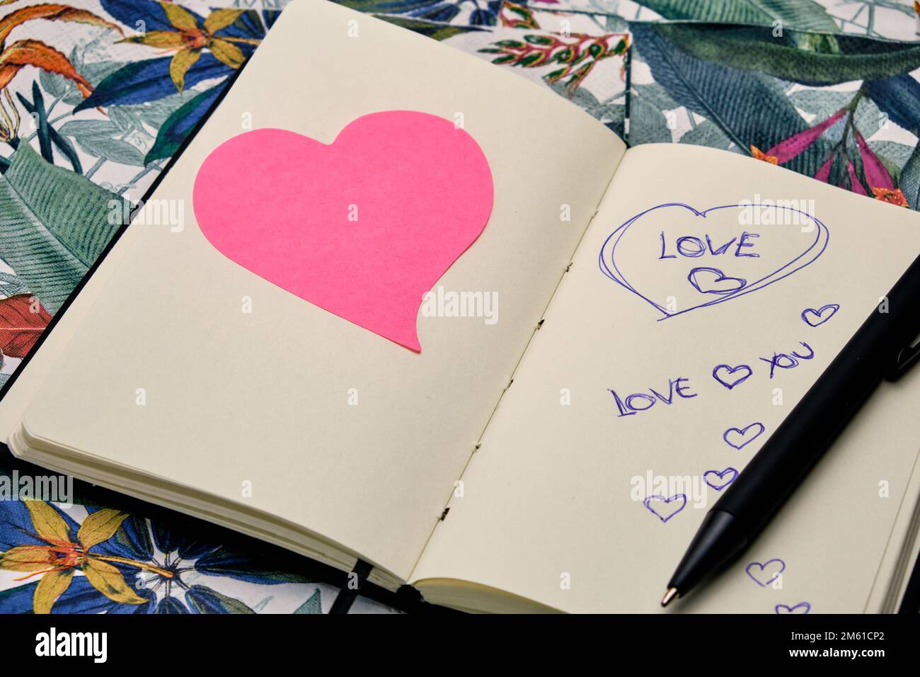 Öffnen Sie das Tagebuch mit einem Stift, einem rosa Herz und schreiben Sie das Wort Liebe auf einen Tisch mit einem tropischen Muster. Stockfoto