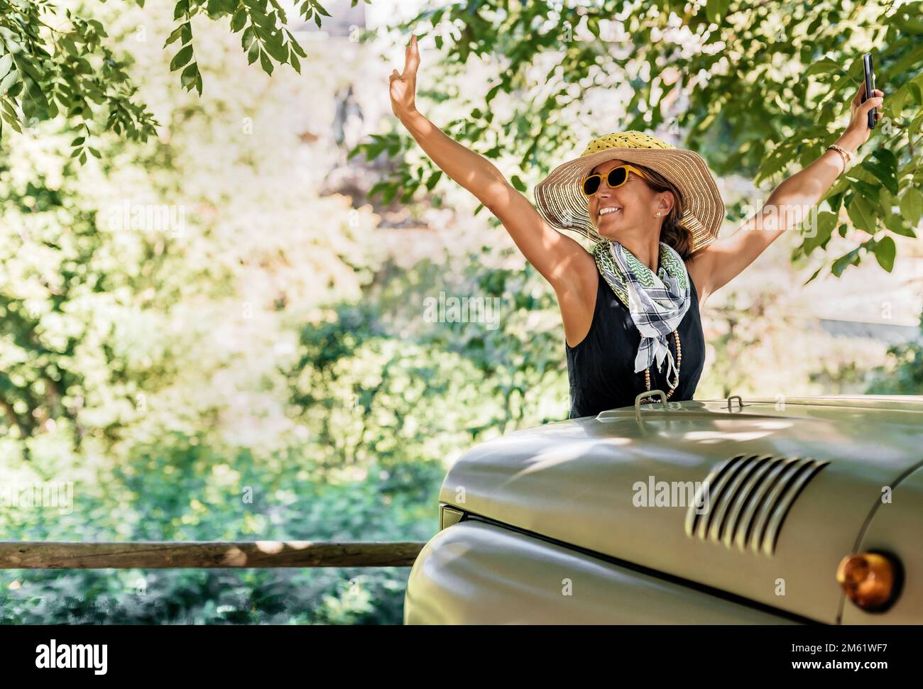 Eine Frau mit Hut und Sonnenbrille hebt die Arme vor einem Geländewagen. Im Hintergrund ein Wald. Konzeptnatur und Pkw-Routen. Stockfoto