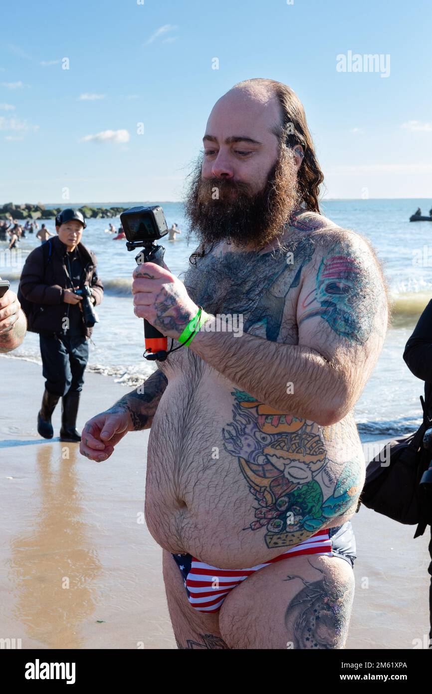 Brooklyn, New York, USA. 1. Januar 2023. Tausende von Badenden und Zuschauern erschienen an einem sonnigen und warmen Neujahrstag für den jährlichen Polarbärsprung auf Coney Island, gesponsert von der Alliance for Coney Island. Ein tätowierter Mann, der einen Slip mit amerikanischer Flagge trägt, filmt nach einem Sprung mit einer Action-Kamera. Kredit: Ed Lefkowicz/Alamy Live News Stockfoto