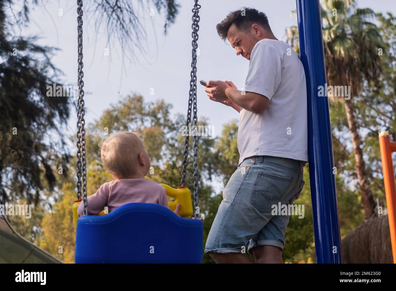 Vater mit Kind auf einem Spielplatz, der auf das Telefon schaut und sein Baby beim Schaukeln ignoriert. Stockfoto