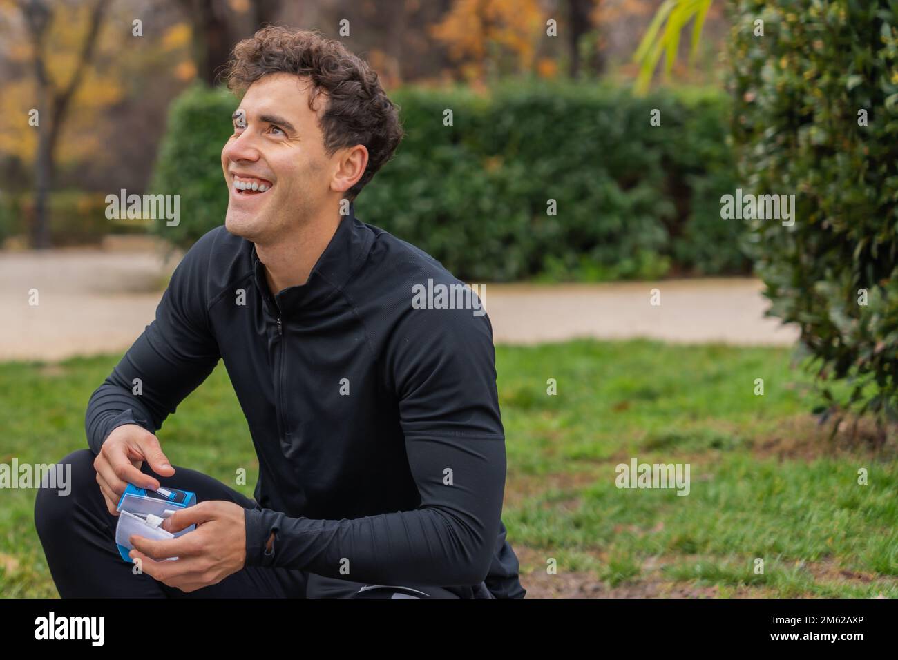 Positiver Mann in lässiger Kleidung, der lächelt und wegschaut, während er auf Gras im grünen Park sitzt Stockfoto