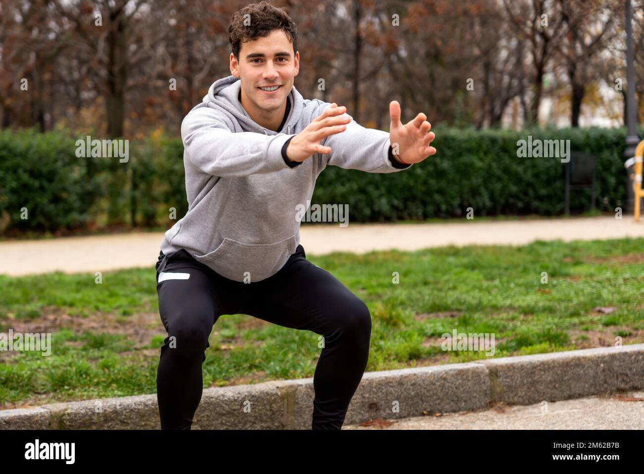 Lächelnder junger Sportler in grauem Hoodie und schwarzen Leggings, der im Park neben grünen Büschen Squats macht und in die Kamera schaut Stockfoto