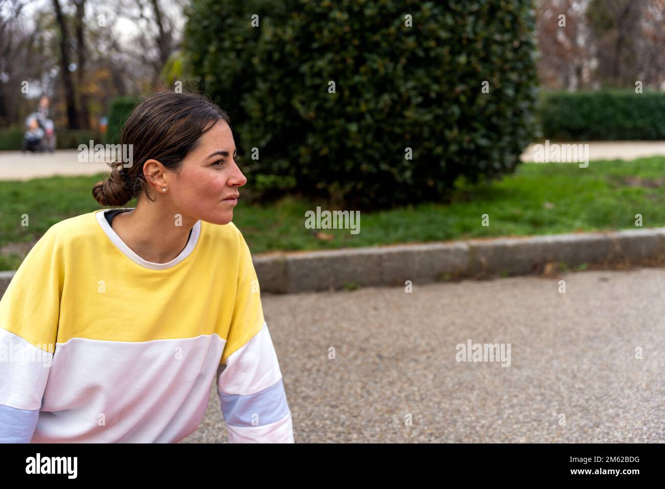 Junge Frau im Sweatshirt blickt aufmerksam weg, während sie im Sommer vor dem unscharfen Hintergrund des grünen Parks sitzt Stockfoto