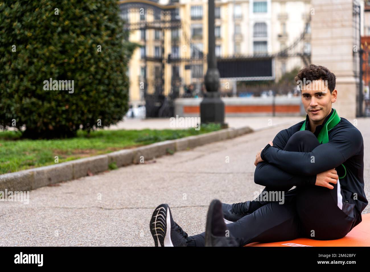 Ein zufriedener, lockiger Typ in Sportbekleidung, der Knie umarmt, während er auf der Straße sitzt und in die Kamera schaut Stockfoto