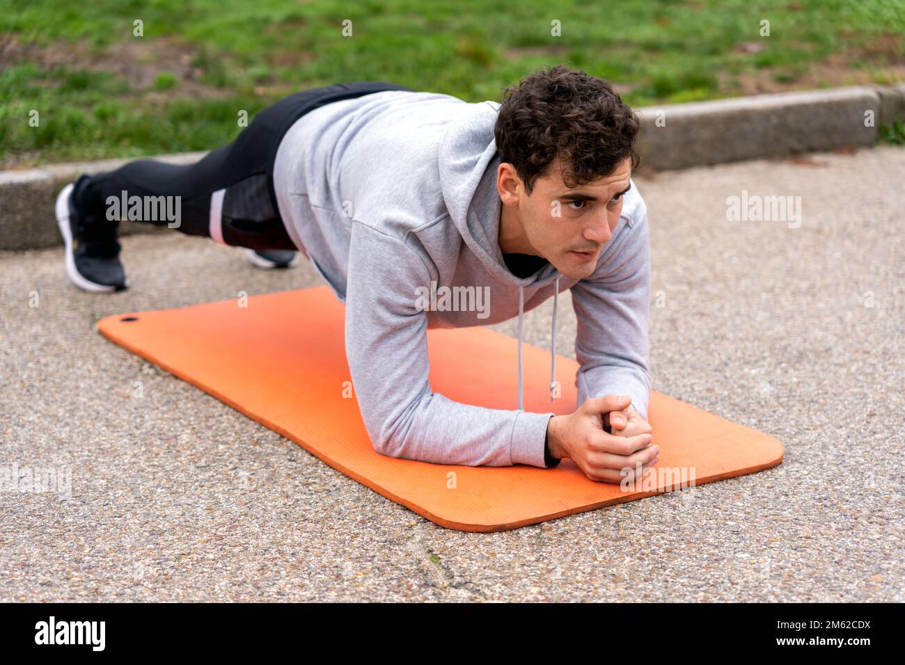 Der ganze Körper eines konzentrierten männlichen Athleten in Aktivbekleidung, der im Park Plank-Training auf einer orangefarbenen Matte macht Stockfoto