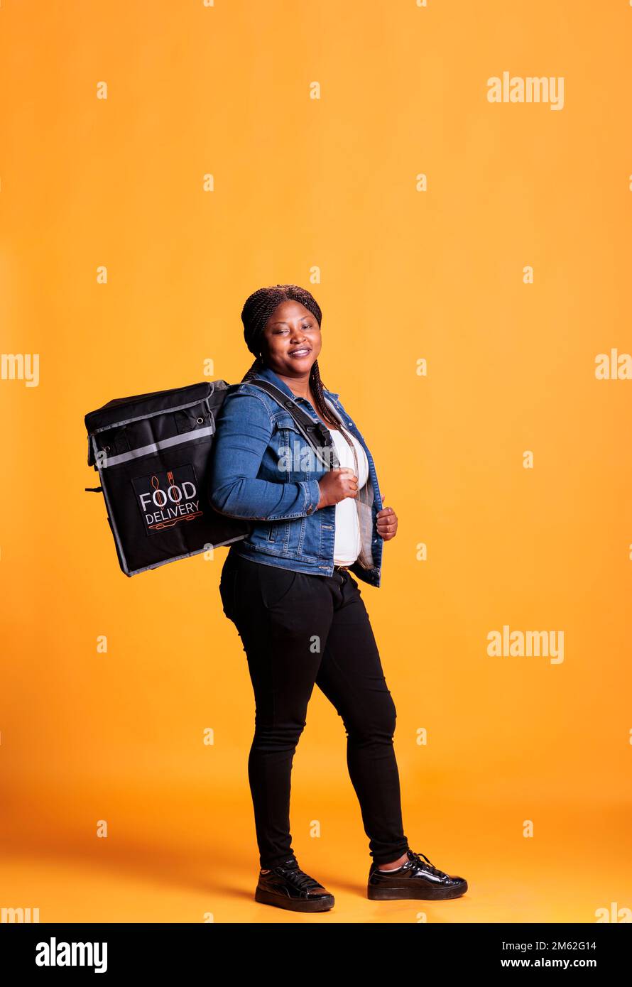 Lieferfrau, die im Studio auf gelbem Hintergrund steht und einen Thermorucksack zum Mitnehmen mitführt, der die Bestellung an den Kunden ausliefert. Gastronomieservice und Mitnahmekonzept Stockfoto