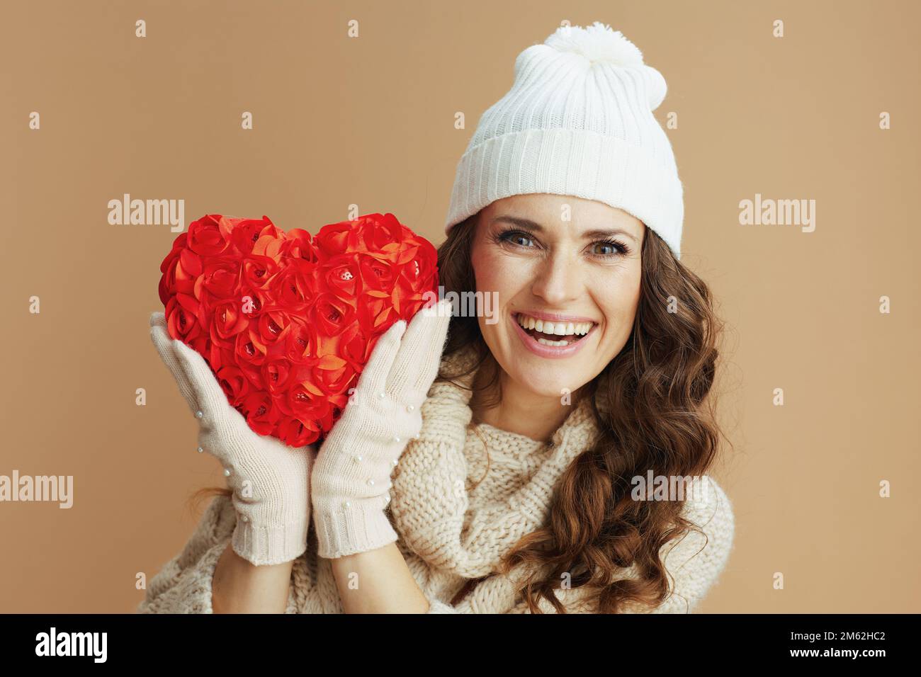 Hallo Winter. Porträt einer glücklichen modernen Frau mittleren Alters in beigefarbenem Pullover, Fäustlingen und Hut mit rotem Herzen isoliert auf beige. Stockfoto