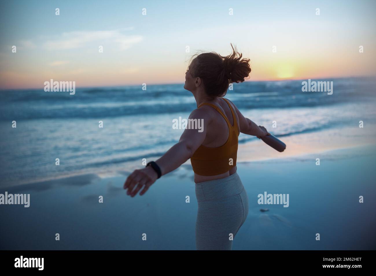 Entspannte, aktive Sportlerin in Fitnesskleidung mit einer Flasche Wasser, die sich am Strand bei Sonnenuntergang freut. Stockfoto