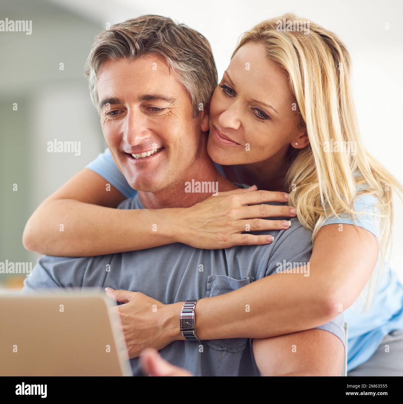 Er vervollständigt mich. Eine attraktive, reife Frau, die ihren hübschen, reifen Ehemann umarmt, während er ein Notebook im Haus benutzt. Stockfoto