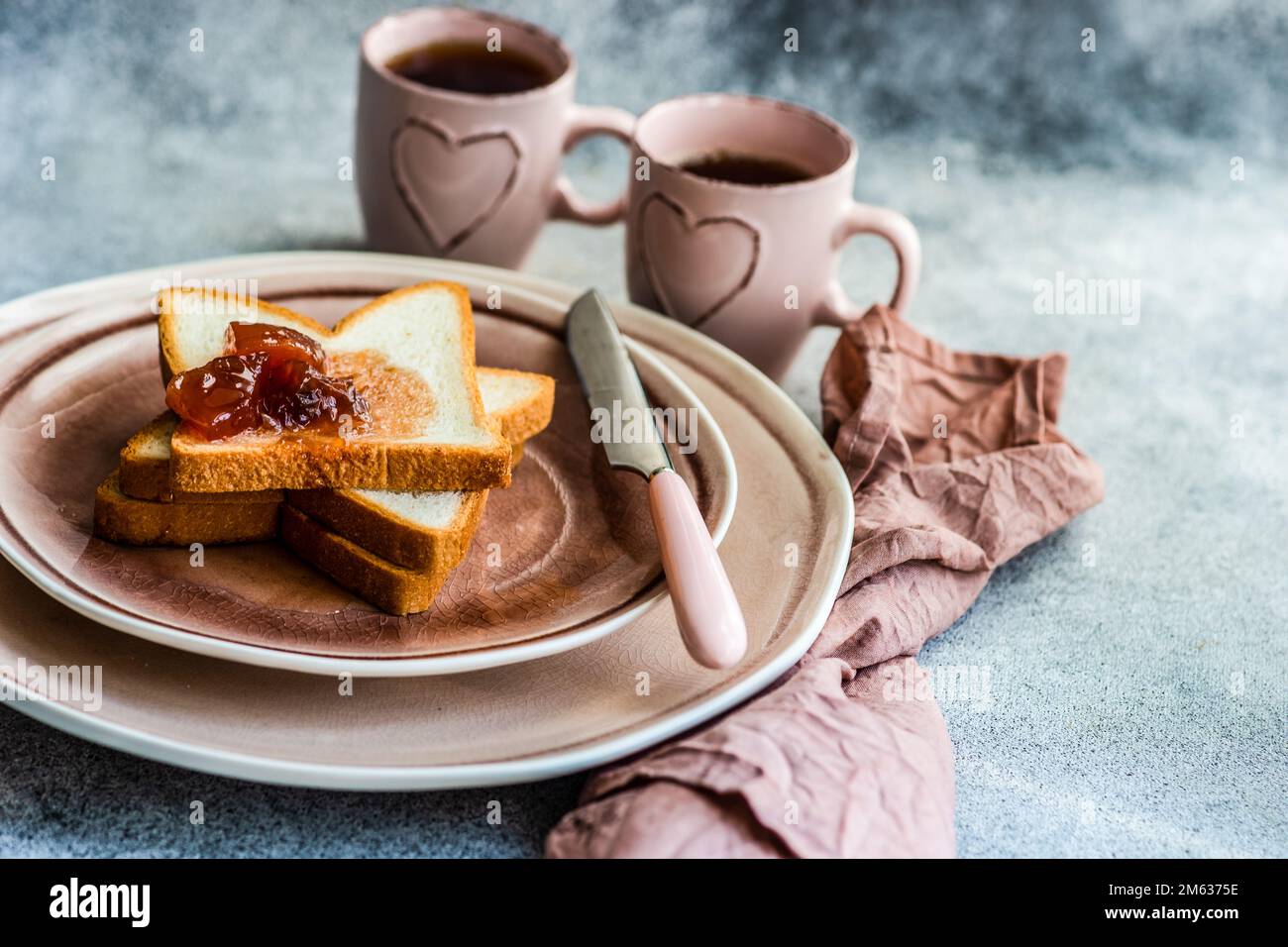 Traditionelles Frühstück mit Marmelade auf Toast auf pinkfarbenen Keramiktellern und einer Tasse Tee auf dem Betontisch Stockfoto