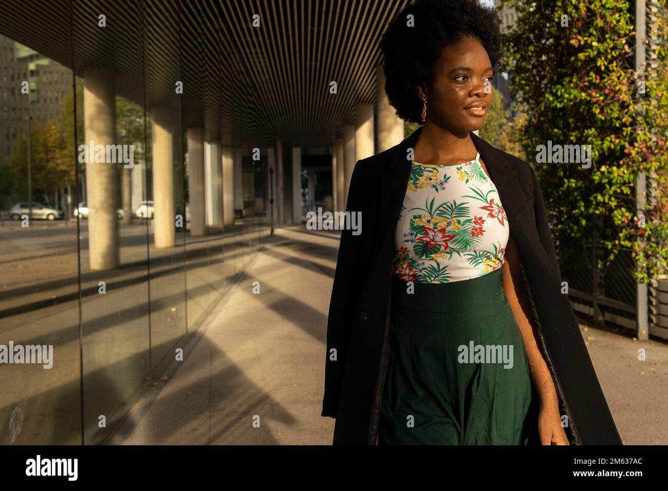 Nachdenkliche junge afroamerikanische Frau in schwarzer Jacke, die neben dem Gebäude steht und an sonnigen Tagen mit Bedacht wegschaut Stockfoto