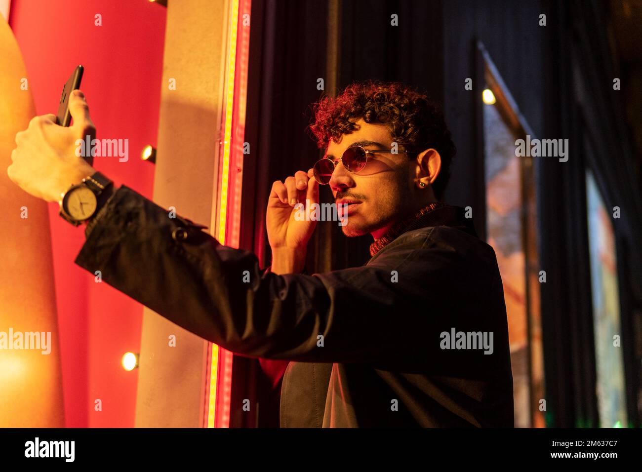 Kleiner Winkel eines jungen Mannes in stilvoller Kleidung, der eine Sonnenbrille anpasst, während er Selfie auf dem Handy macht, wenn er auf der Straße steht Stockfoto