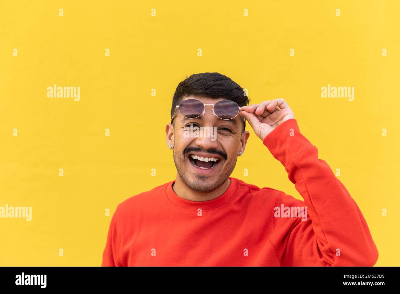 Glücklicher junger spanischer Mann in rotem Sweatshirt, der eine stilvolle Sonnenbrille hebt und mit einem Lächeln auf gelbem Hintergrund in die Kamera schaut Stockfoto