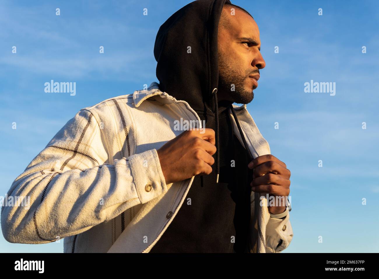 Ein männlicher Afroamerikaner in Jacke und Kapuze mit Kapuze auf dem Kopf, der auf der Straße gegen Sonnenlicht läuft und wegschaut Stockfoto