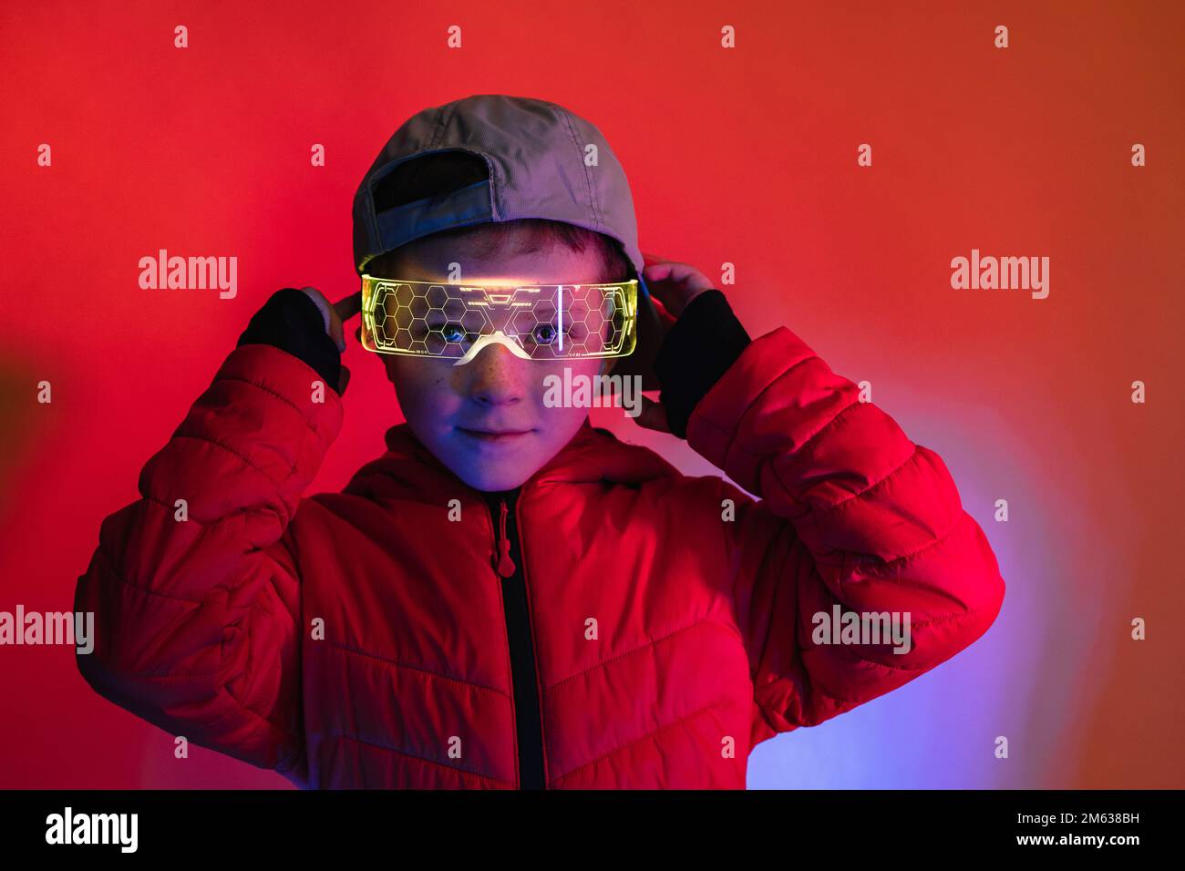 Ein Junge mit blauem Mantel und Kappe, der die leuchtende digitale Brille anpasst und unter roter Neonbeleuchtung in die Kamera schaut Stockfoto