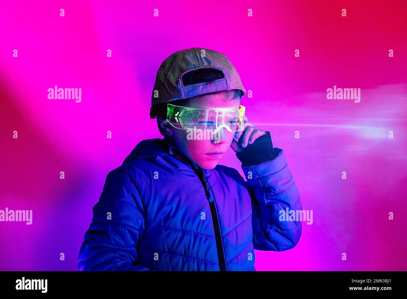 Der Junge mit blauem Mantel und Kappe passt die leuchtende digitale Brille an und blickt unter magentafarbener Neonbeleuchtung weg Stockfoto