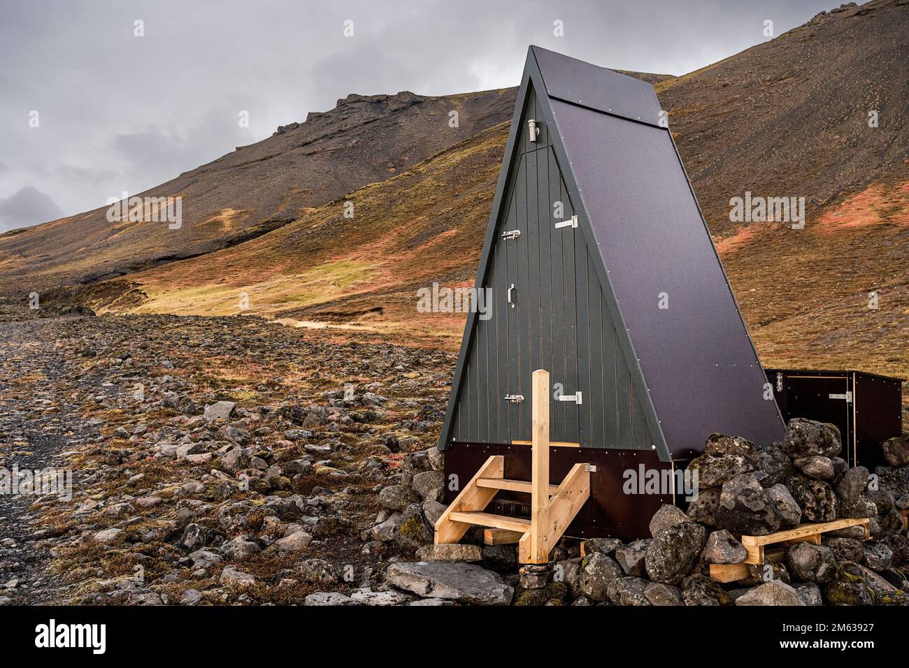 Dreieckige hölzerne, lange Toilettenkabine auf rauem felsigem Gelände in der Nähe vulkanischer Berge unter grauem, wolkigen Himmel in Island Stockfoto