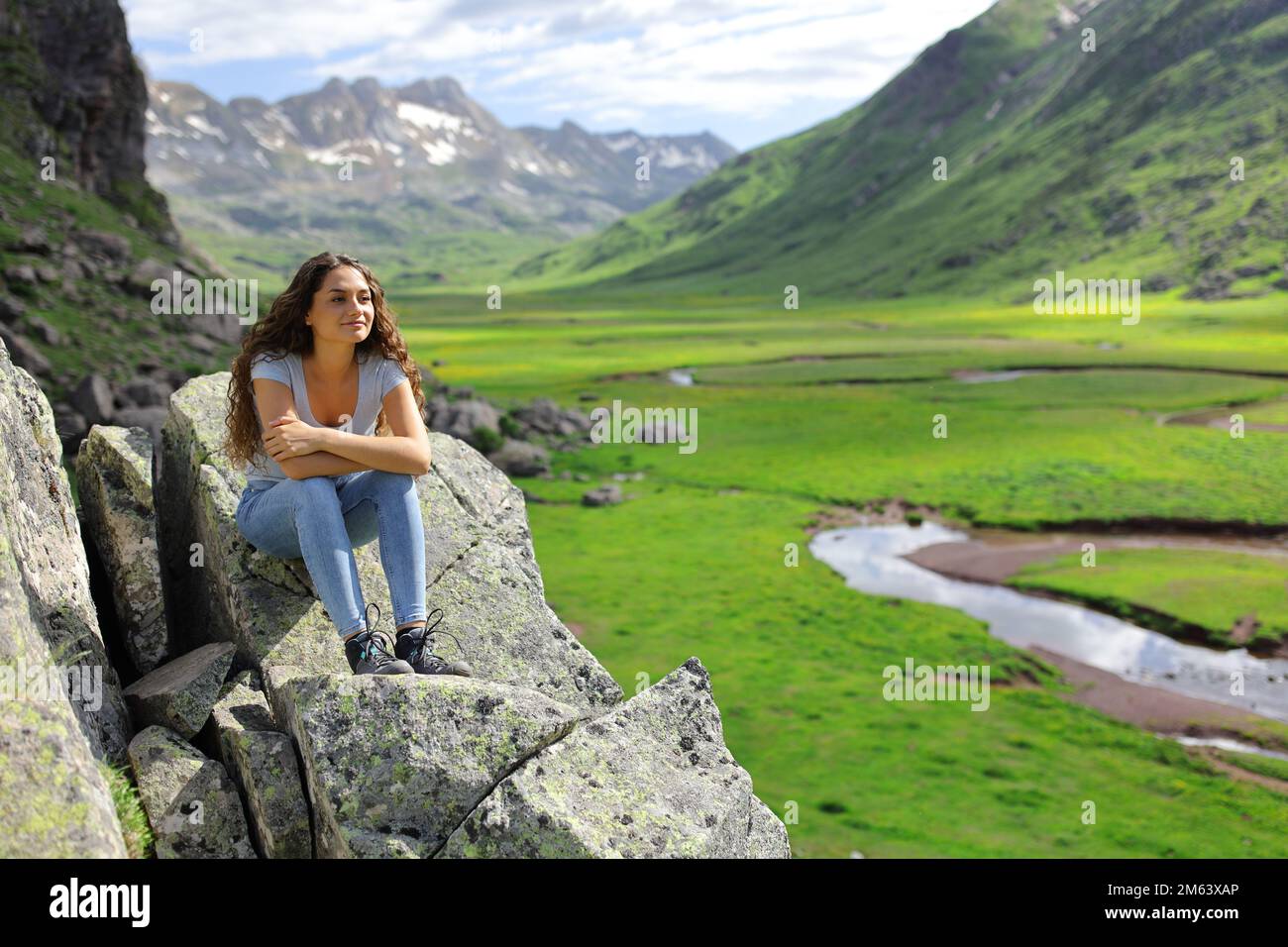 Eine glückliche Frau, die auf einer Klippe sitzt und eine wunderschöne Aussicht auf die Berge in Betracht zieht Stockfoto
