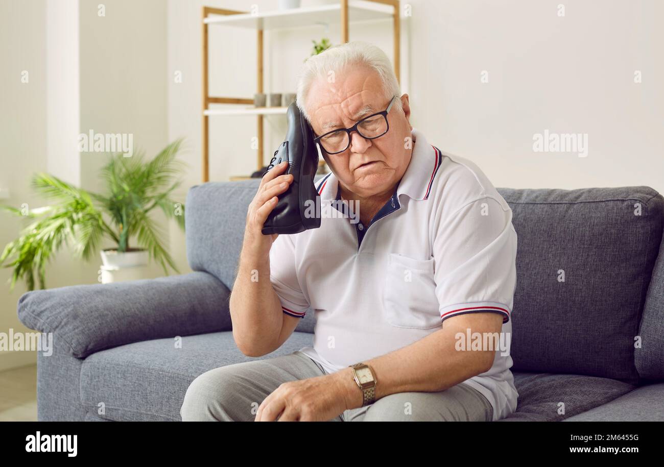 Älterer Mann mit Demenz, der auf dem Sofa sitzt und einen Schuh wie ein Telefon hält Stockfoto