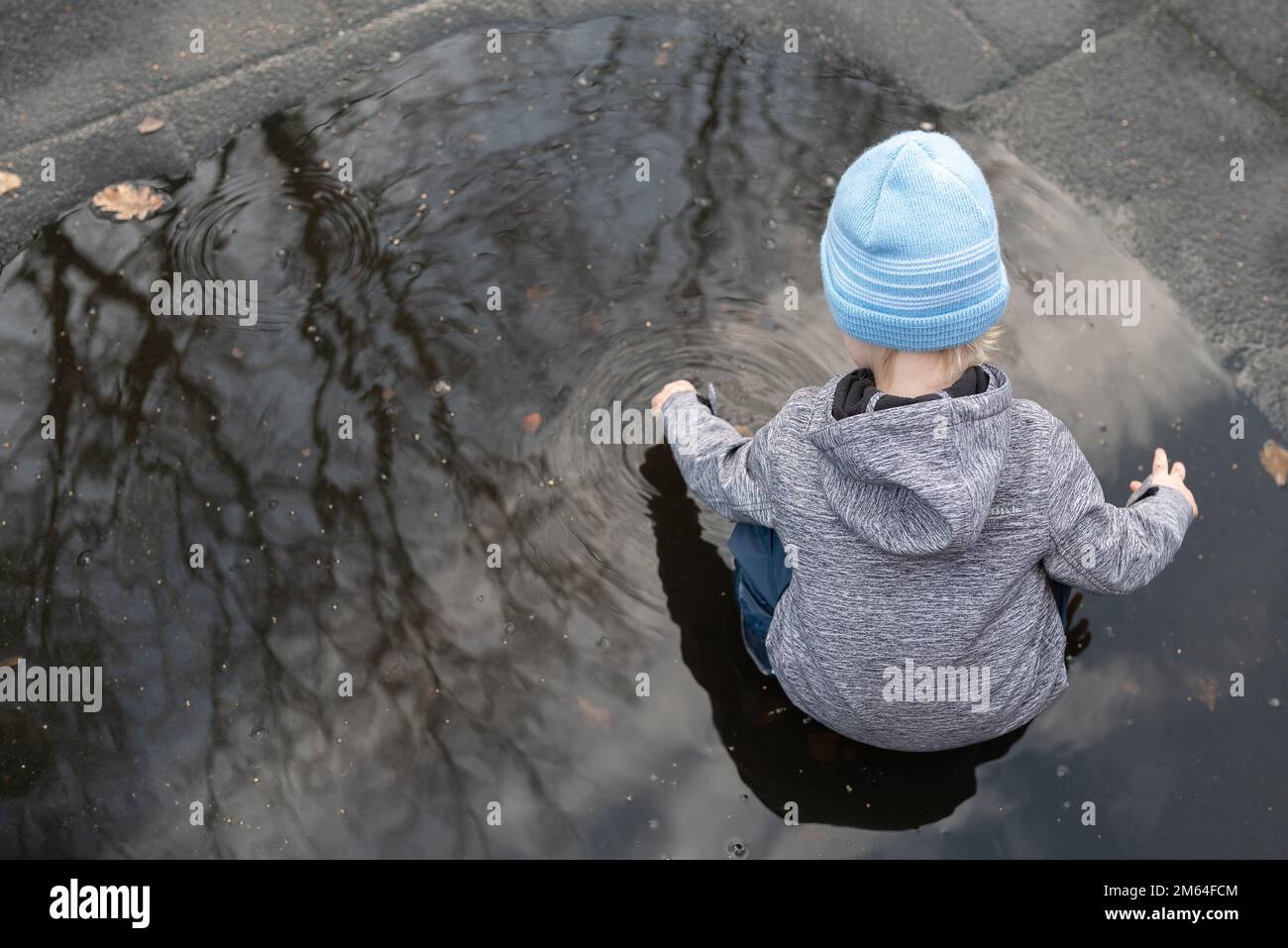 Blick aus einem hohen Winkel auf einen zweijährigen Jungen, der nach einer Regendusche in einer Wasserpfütze spielt Stockfoto